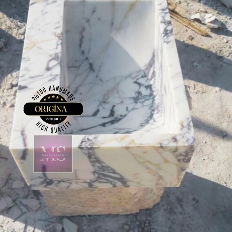 May include: A white marble sink with a square shape and a raised rim. The sink is made of natural stone with a veined pattern. The sink is labeled "100% Handmade Original Product High Quality".