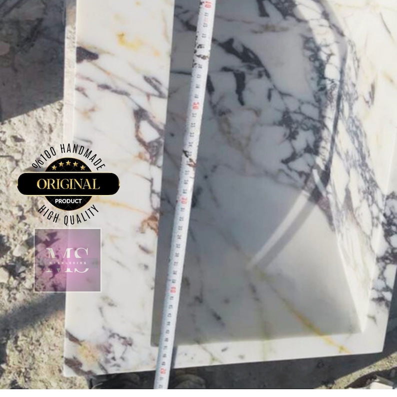 May include: A white marble sink with a rectangular shape and a rounded edge. The sink is surrounded by a white marble countertop with gray and brown veins. A measuring tape is visible on the right side of the image. The sink is labeled "Original Product 100% Handmade High Quality".
