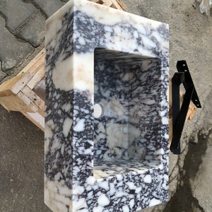 May include: A white marble sink with a black and white veined pattern. The sink is rectangular and has a square cutout for the basin. The sink is sitting on a wooden platform.