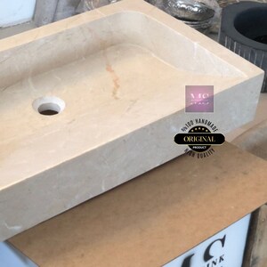 May include: A rectangular, off-white marble sink with a single drain hole. The sink is handmade and features the text "100% Handmade Original Product High Quality" in a circle.
