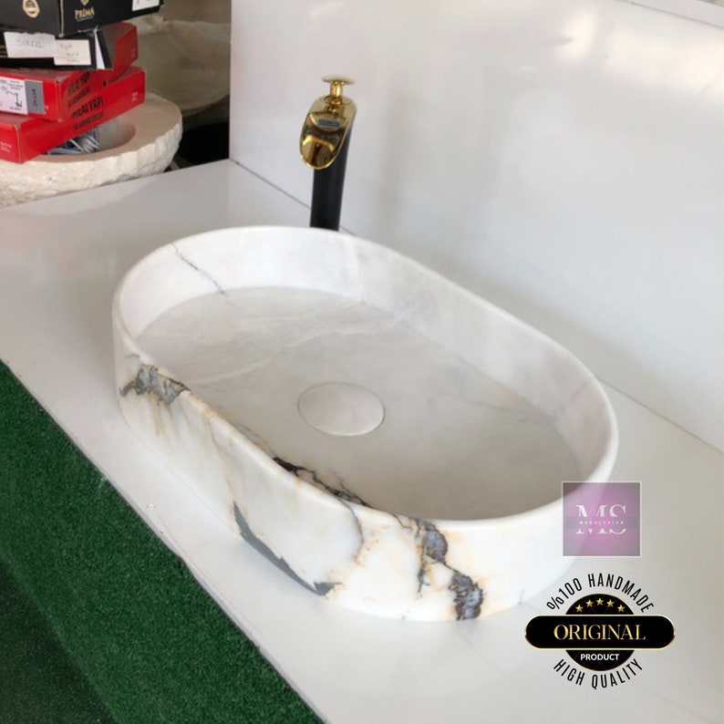 May include: White oval marble sink with a gold faucet and a black base. The sink has a drain in the center and is surrounded by a white countertop. The sink is labeled "100% Handmade Original Product High Quality".