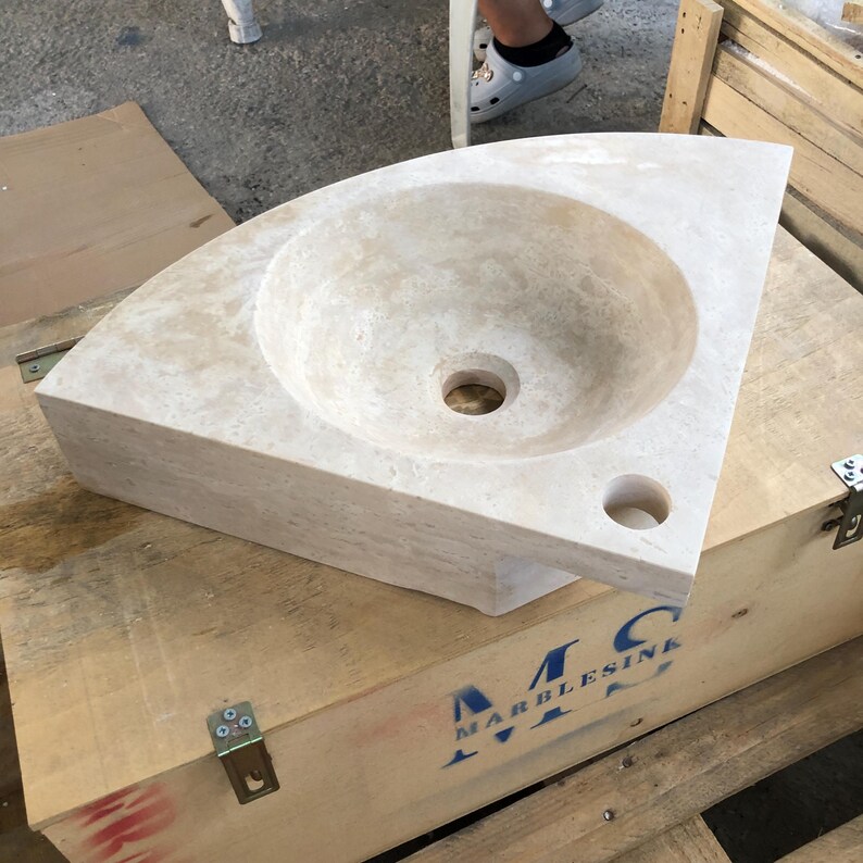 May include: A corner sink made of light beige travertine stone. The sink is in a wooden crate with the text "MARBLESINA" printed on the side.
