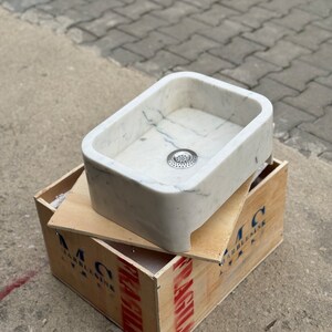 High-angle front view of a Carrara White marble vessel sink showing the rounded rectangular bowl, centered drain opening, thick stone rim and natural grey veining.