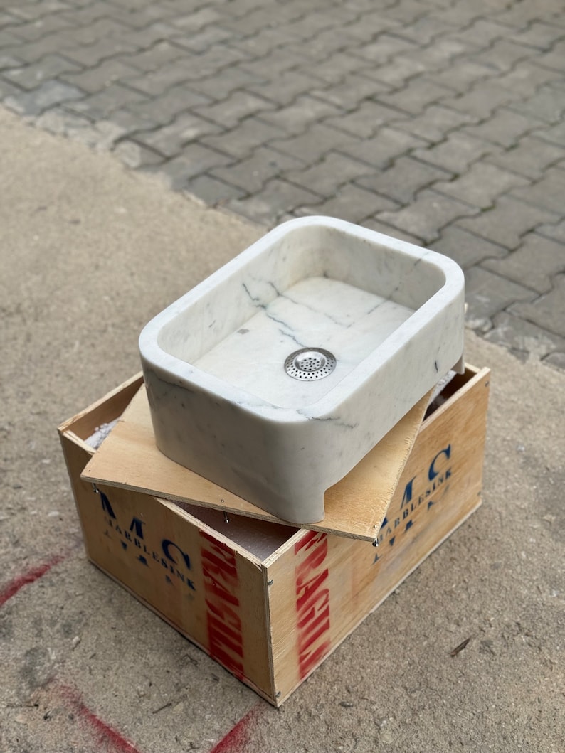 High-angle front view of a Carrara White marble vessel sink showing the rounded rectangular bowl, centered drain opening, thick stone rim and natural grey veining.