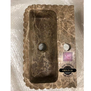 May include: A brown marble bathroom sink with a wavy edge and two holes. The sink is handmade and has a high quality finish. The text "Original Product" is visible on a sticker.