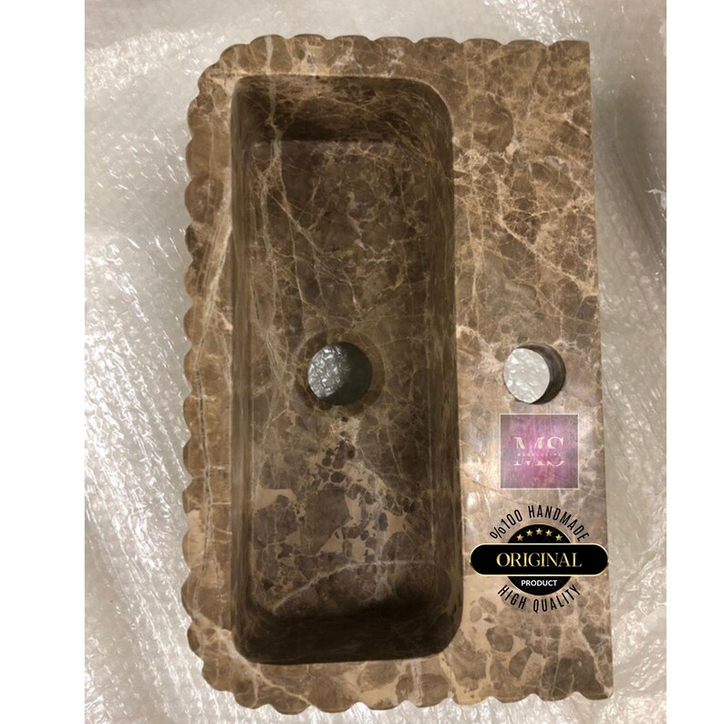 May include: A brown marble bathroom sink with a wavy edge and two holes. The sink is handmade and has a high quality finish. The text "Original Product" is visible on a sticker.