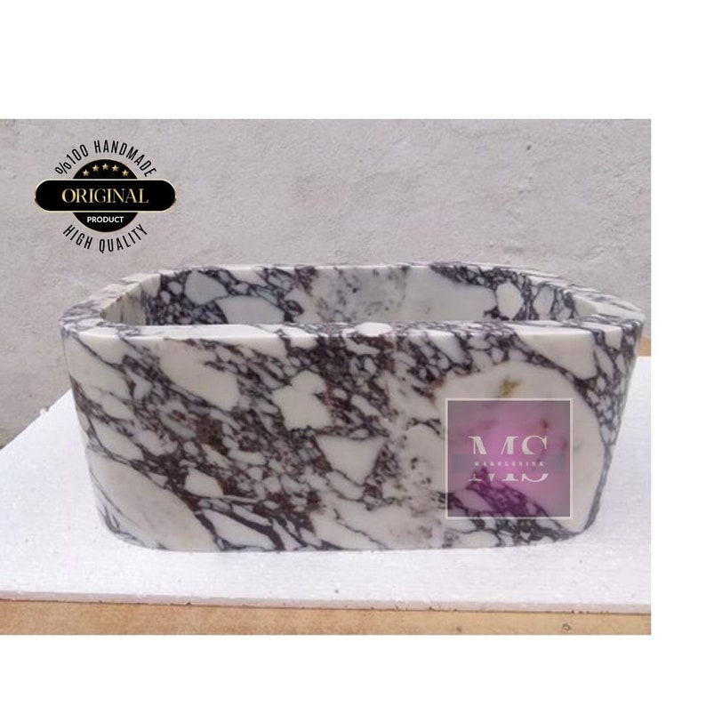 Op de afbeelding: Een witte marmeren gootsteen met paarse en grijze aderen. De gootsteen is rechthoekig met afgeronde randen en is handgemaakt. De gootsteen is gelabeld met de tekst "MC Marble Creations".