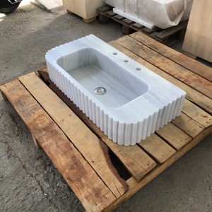May include: A white marble sink with a fluted edge sits on a wooden pallet. The sink has a single drain and two small holes for taps.