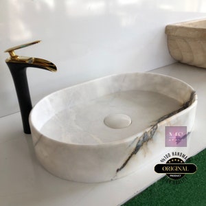 May include: White oval marble sink with a black and gold faucet. The sink has a drain in the center and a subtle gray veining pattern. The text "ORIGINAL PRODUCT HIGH QUALITY" is visible on a sticker on the sink.
