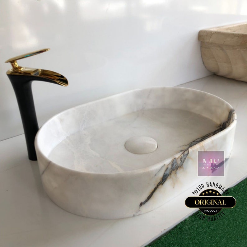 May include: White oval marble sink with a black and gold faucet. The sink has a drain in the center and a subtle gray veining pattern. The text "ORIGINAL PRODUCT HIGH QUALITY" is visible on a sticker on the sink.