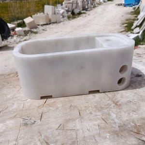May include: A white marble bathtub with an oval shape. The tub has a rectangular top with a small, raised section and two circular holes on one side. The base has four rectangular cutouts. The tub is sitting on a wooden surface.