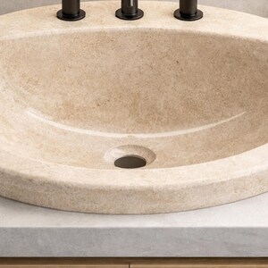 May include: A beige oval bathroom sink with a textured surface. The sink has a dark drain and three dark faucet handles. The sink is set on a light gray countertop.