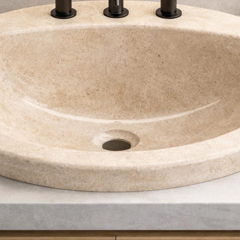 May include: A beige oval bathroom sink with a textured surface. The sink has a dark drain and three dark faucet handles. The sink is set on a light gray countertop.