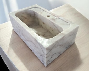 May include: A white marble bathroom sink with a rectangular shape and a rounded interior. The sink is sitting on a light wood surface.