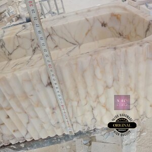 May include: A white marble bathtub with a rippled edge. The tub is 29 inches wide and is being constructed. The text "96100 HANDMADE ORIGINAL PRODUCT HIGH QUALITY" is visible on the side of the tub.