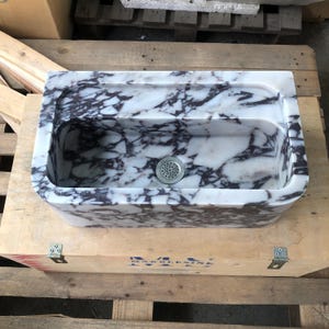 May include: A white marble sink with purple veining. The sink has a rounded rectangular shape and a single drain hole. The sink is sitting on a wooden crate.