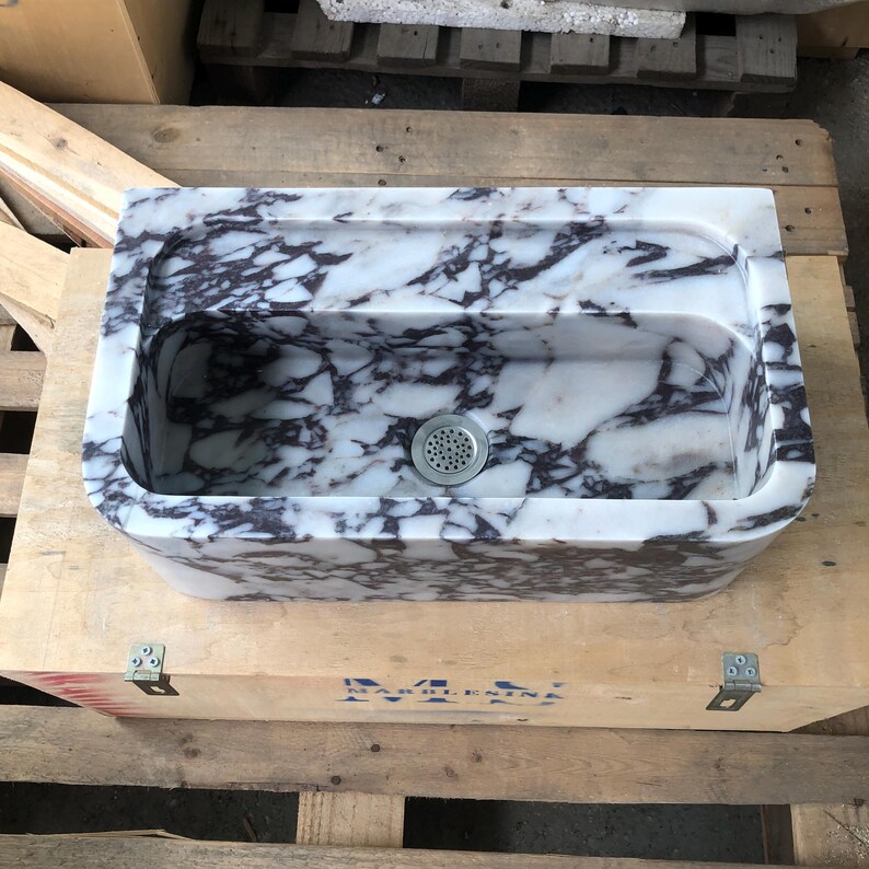 May include: A white marble sink with purple veining. The sink has a rounded rectangular shape and a single drain hole. The sink is sitting on a wooden crate.