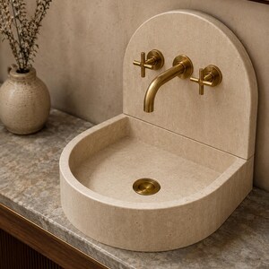May include: A beige stone sink with a rounded back panel and a curved basin. The sink features a wall-mounted brass faucet with two cross-handle controls and a matching brass drain cover, creating a cohesive look for a bathroom vanity setup.