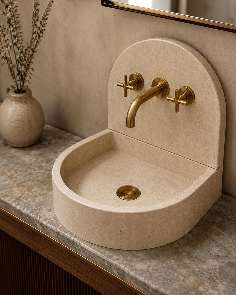May include: A beige stone sink with a rounded back panel and a curved basin. The sink features a wall-mounted brass faucet with two cross-handle controls and a matching brass drain cover, creating a cohesive look for a bathroom vanity setup.