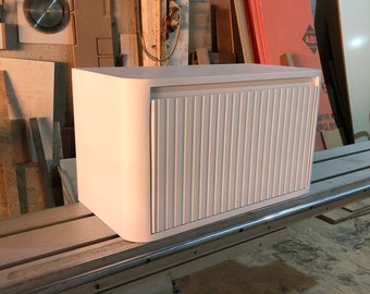 May include: White cabinet with a rounded edge and a slatted door. The cabinet is made of wood and has a modern design.