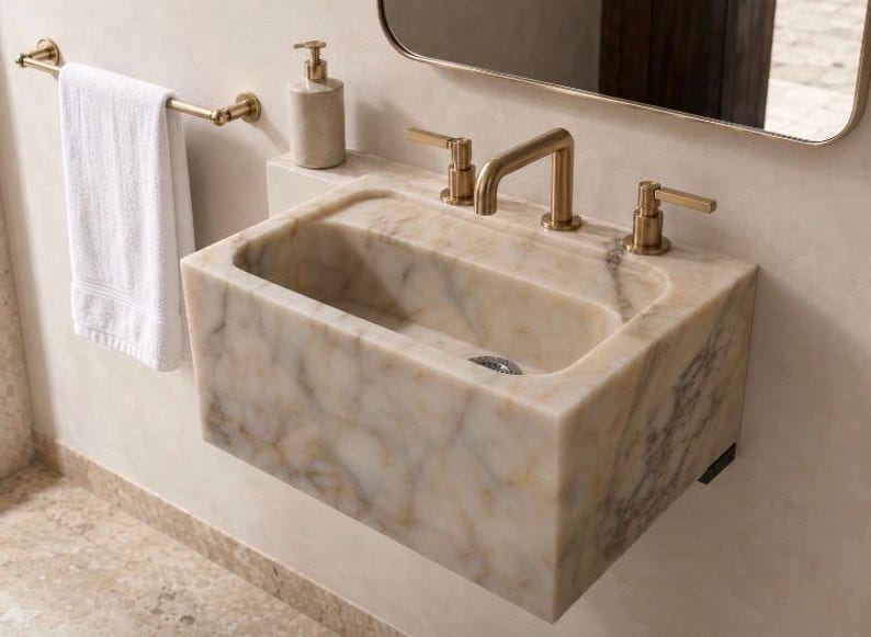 Wall-mounted Calacatta Viola marble sink installed in a modern powder room with brushed brass three-hole faucet, rounded mirror and white towel, showing the deep rectangular basin and natural cream veining.