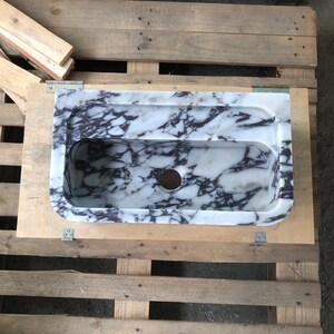 May include: A white marble sink with purple veining. The sink has a rounded rectangular shape and a single drain hole.