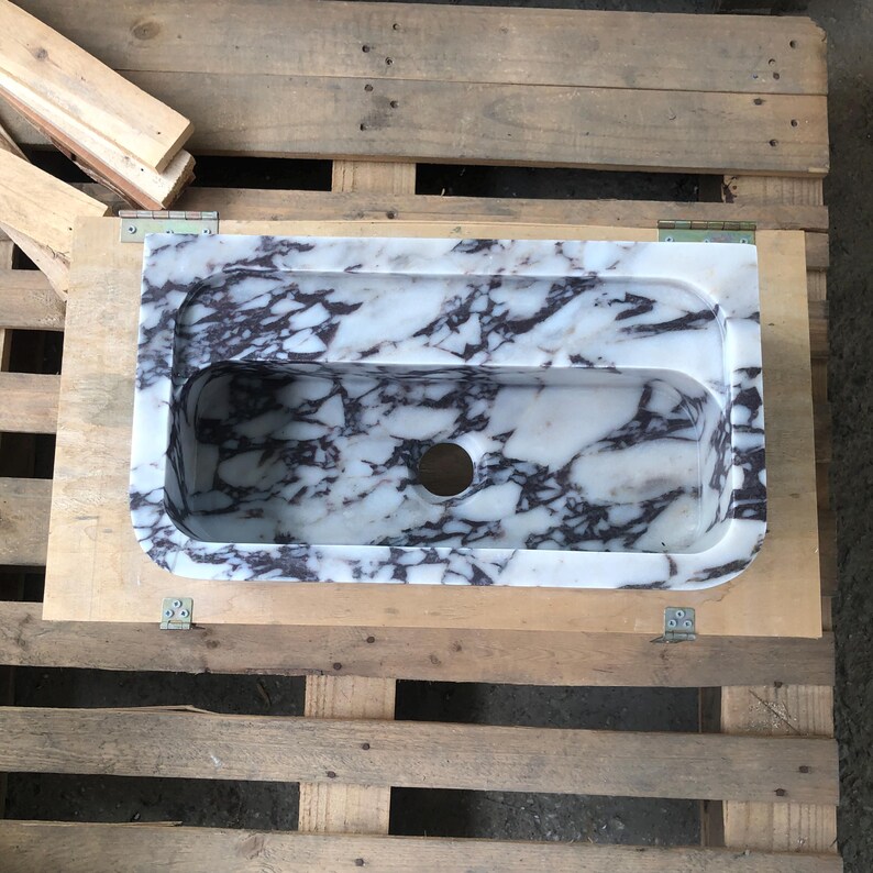 May include: A white marble sink with purple veining. The sink has a rounded rectangular shape and a single drain hole.