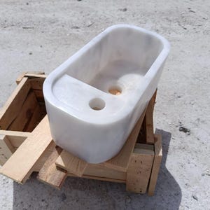 May include: A rectangular white marble sink with two circular drain holes. The sink is set on a wooden pallet. The sink has a smooth, polished surface and is designed for bathroom use.