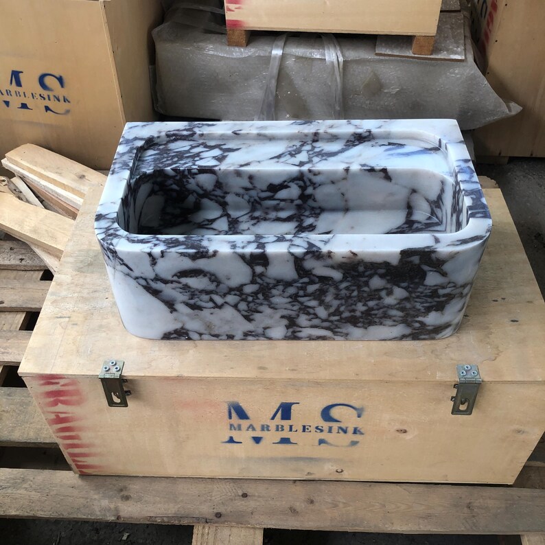 May include: A white marble sink with purple veining. The sink is in a wooden crate with the text "MC MARBLESINK" printed on the side.