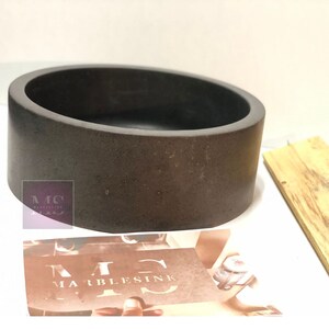 Pu&ograve; includere: Un lavabo rotondo in cemento grigio scuro con una superficie liscia leggermente strutturata. Il lavabo &egrave; progettato per un bagno o un bagno di cortesia.