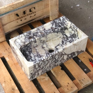 Puede incluir: Un lavabo de m&aacute;rmol blanco con vetas moradas y grises. El lavabo tiene dos orificios de drenaje y es rectangular.