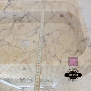 May include: A white marble sink with a wavy edge and a drain hole. The sink is 47 inches long and is being measured with a tape measure. The sink is handmade and has a high quality finish.