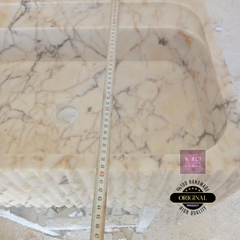 May include: A white marble sink with a wavy edge and a drain hole. The sink is 47 inches long and is being measured with a tape measure. The sink is handmade and has a high quality finish.