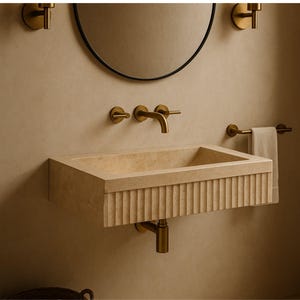 May include: A beige rectangular bathroom sink with a fluted front, paired with a brass faucet and fixtures. A round black mirror and a towel bar with a beige towel are also visible. The sink is mounted on a beige wall.