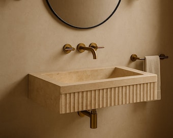 May include: A beige rectangular bathroom sink with a fluted front, paired with a brass faucet and fixtures. A round black mirror and a towel bar with a beige towel are also visible. The sink is mounted on a beige wall.