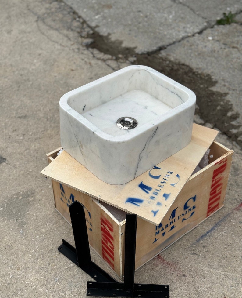 Right three-quarter workshop view of a Carrara White marble bathroom sink showing its thick solid stone walls, rounded corners, centered drain and black wall-mount brackets.