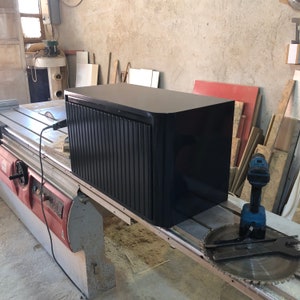 May include: A black cabinet with a slatted front door. The cabinet is sitting on a workbench in a workshop.