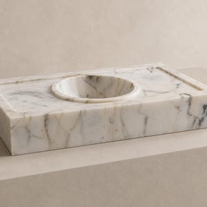 Hand carved Calacatta Gold marble sink installed as a floating wall mounted vanity with brushed brass faucet, dark gray luxury bathroom background and sculptural accessories