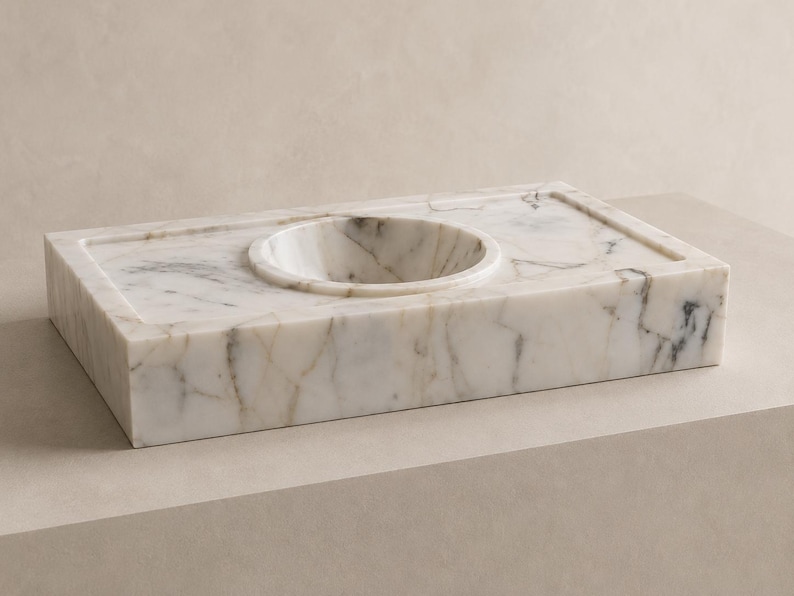 Hand carved Calacatta Gold marble sink installed as a floating wall mounted vanity with brushed brass faucet, dark gray luxury bathroom background and sculptural accessories