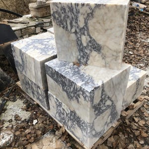 May include: Four blocks of white marble with gray and purple veining stacked on top of each other. The blocks are sitting on a wooden pallet.