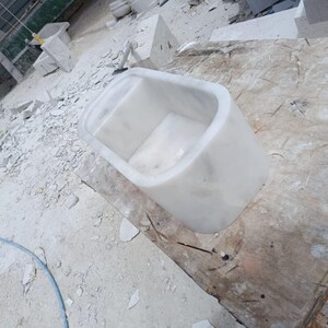 May include: A white marble bathtub with rounded edges. The tub is rectangular with a smooth, polished finish. It sits on a wooden surface in a workshop setting, surrounded by marble debris.