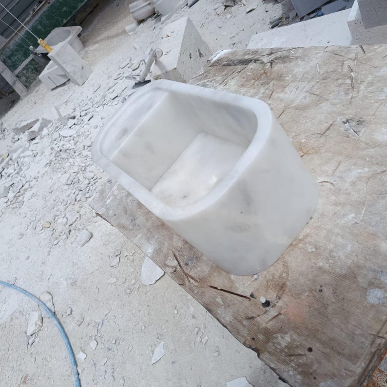 May include: A white marble bathtub with rounded edges. The tub is rectangular with a smooth, polished finish. It sits on a wooden surface in a workshop setting, surrounded by marble debris.