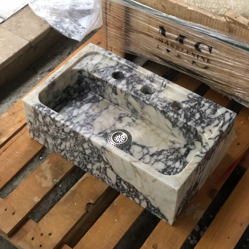 Puede incluir: Un lavabo de m&aacute;rmol blanco con vetas moradas y grises. El lavabo tiene tres agujeros para grifos y un desag&uuml;e en el centro. El lavabo est&aacute; sobre una tarima de madera.