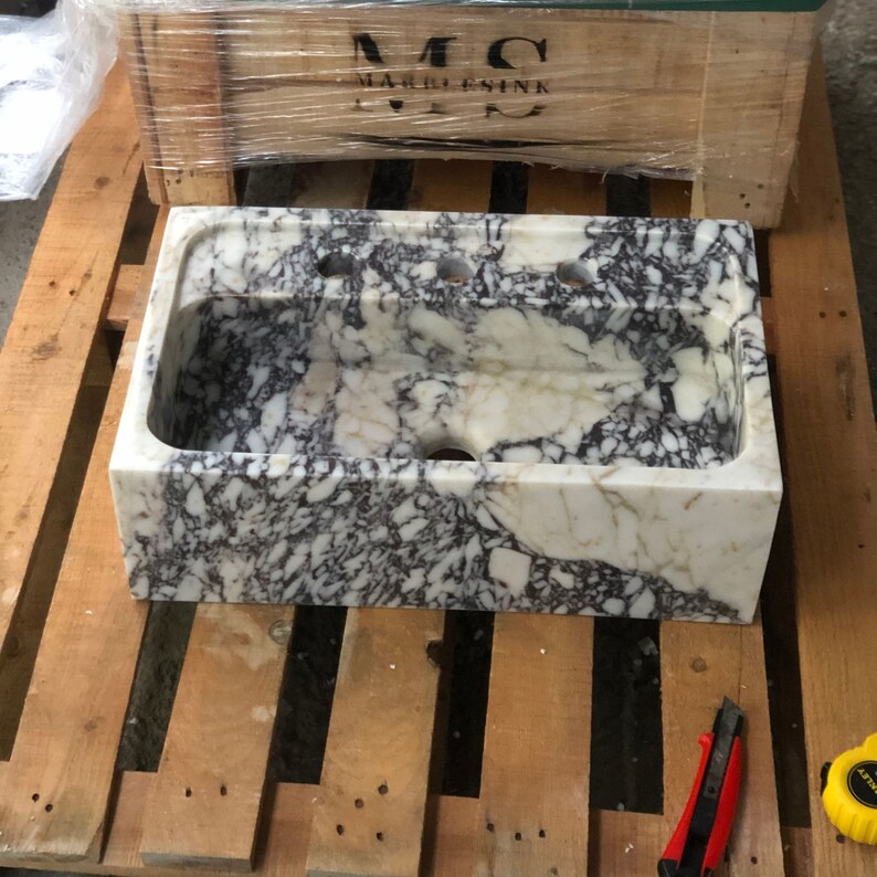 Puede incluir: Un lavabo de m&aacute;rmol blanco con vetas negras. El lavabo tiene cuatro agujeros para grifos y un agujero de desag&uuml;e. El lavabo est&aacute; sentado en una plataforma de madera.