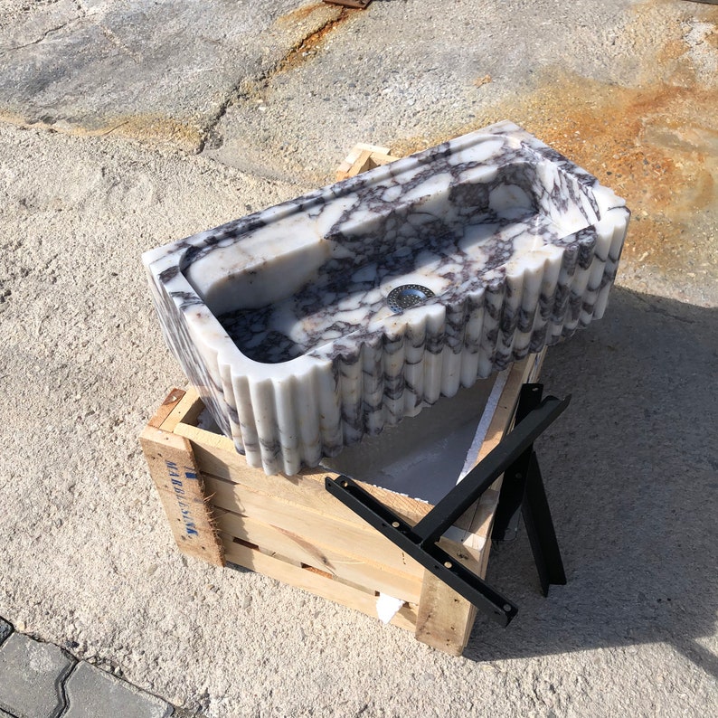Three-quarter workshop view of the handmade fluted marble basin with black wall-mounting supports beside the wooden crate.