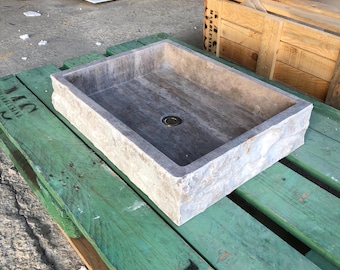 Peut inclure: Un évier rectangulaire en pierre avec un extérieur rugueux et texturé. L'évier est fait d'une pierre brun clair avec des veines brun foncé. L'évier est posé sur une palette en bois vert.