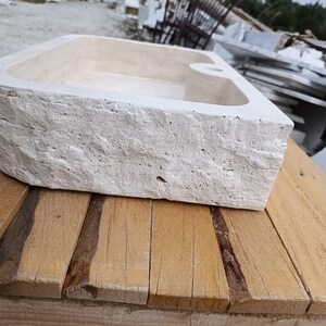 Op de afbeelding: Een rechthoekige stenen wastafel met een ruwe, gebeitelde buitenkant en een gladde, lichtbeige binnenkant. De wastafel heeft een platte rand met een enkel rond gat voor een kraan, wat de natuurlijke poreuze steendetails benadrukt.