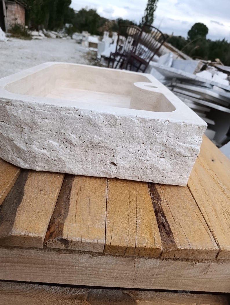 Op de afbeelding: Een rechthoekige stenen wastafel met een ruwe, gebeitelde buitenkant en een gladde, lichtbeige binnenkant. De wastafel heeft een platte rand met een enkel rond gat voor een kraan, wat de natuurlijke poreuze steendetails benadrukt.
