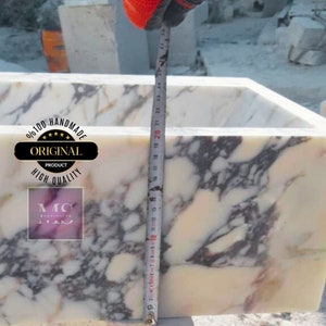 May include: A white marble sink with pink and grey veining. The sink is 20 inches tall and is being measured with a tape measure. The sink is handmade and is labeled as an original product.