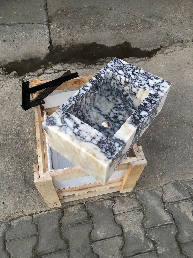 May include: A white marble sink with black and gray veining, in a wooden crate. The sink is rectangular and has a rounded edge. The sink has a drain hole in the center.